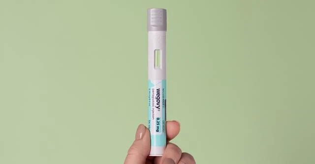 A hand holding a single injector pen against a plain sage-green background.